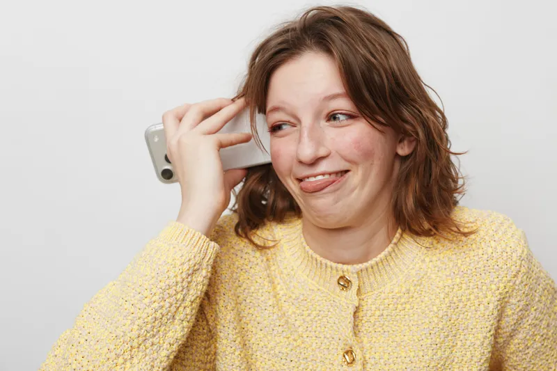 Young woman in yellow sweater makes funny face while holding smartphone near ear. Casual lifestyle, communication, humor, and playful mood on white background.