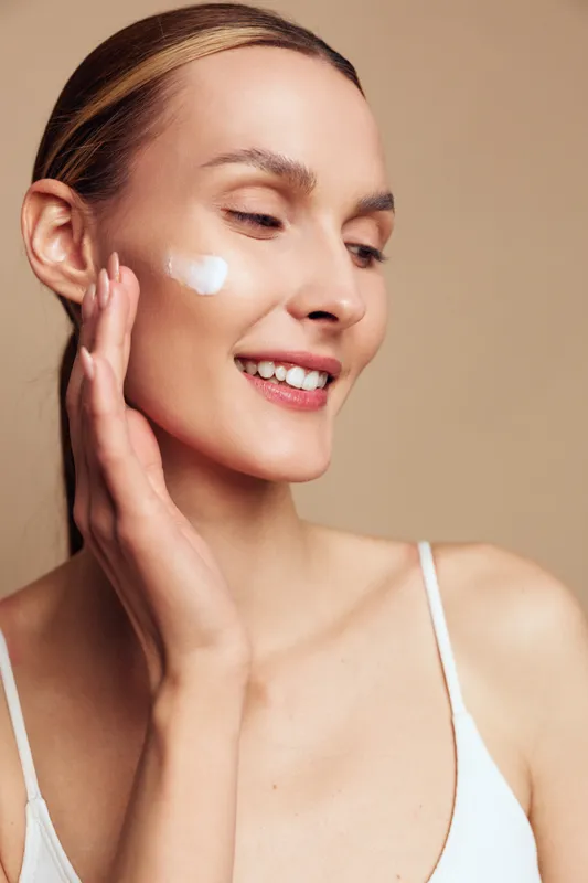 Young woman with clear skin applying white face cream on cheek. Close-up portrait with soft lighting on beige background. Concept of skincare, beauty, and healthy lifestyle.