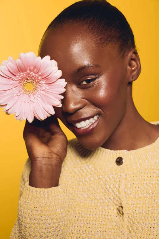 Smiling black woman with dark skin holds pink flower near face against yellow background. Portrait of happy African female model in yellow sweater. Concept of beauty, nature, and positive mood.