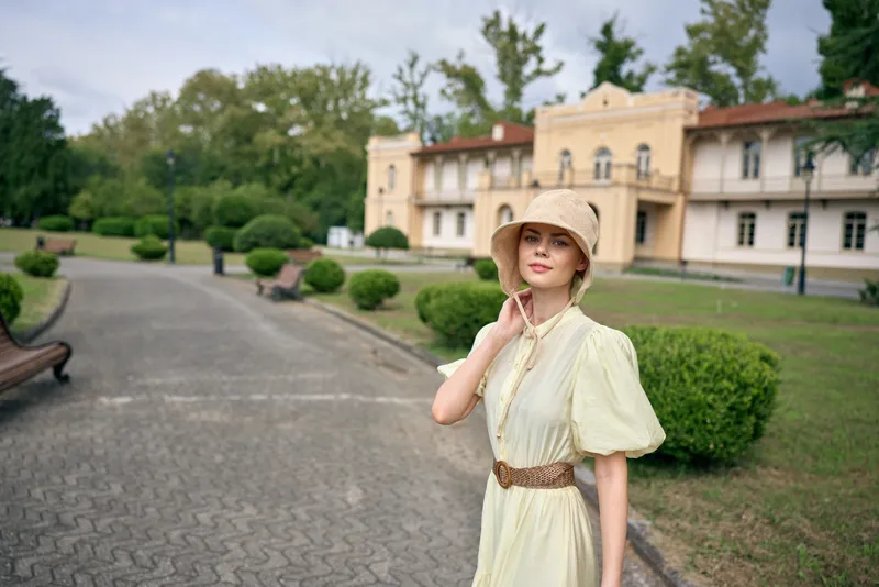 Woman in a vintage dress and wide hat stands outdoors in a manicured garden with an elegant estate in the background, creating a timeless fashion portrait