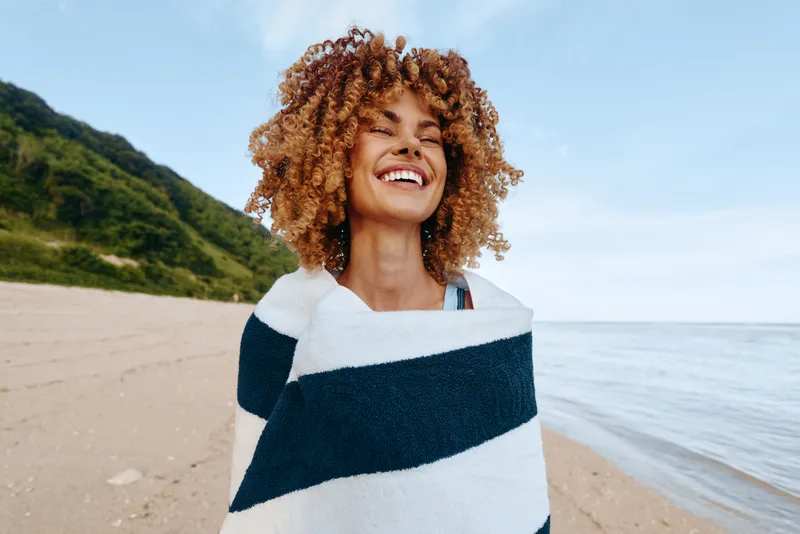 A smiling woman on a sunny beach wrapped in a striped towel, enjoying the breeze. Relaxed outdoor portrait conveying warmth, leisure, and carefree summer mood.