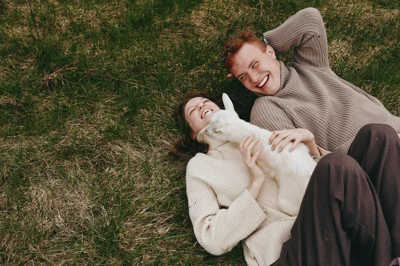 Young couple lying on grass with a white baby goat enjoying a playful moment together in cozy sweaters during a casual lifestyle outdoor scene.