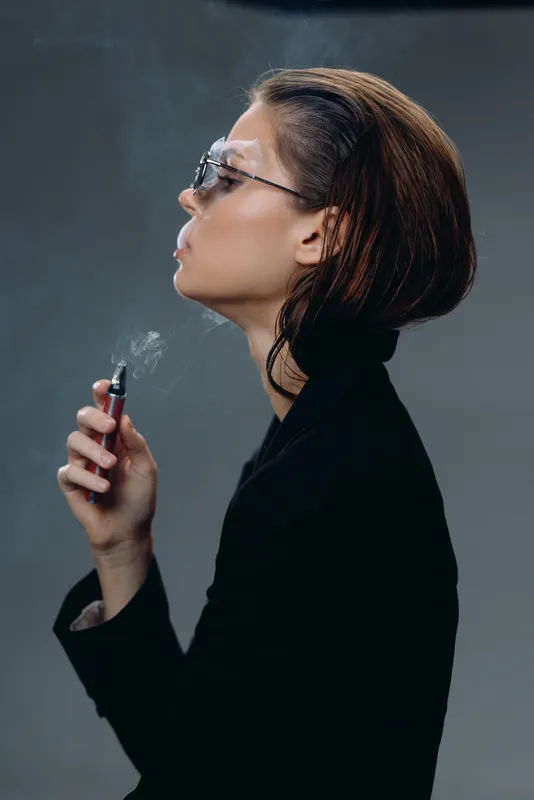 Profile of a stylish woman with glasses smoking electronic cigarette in dark clothing and smoky background