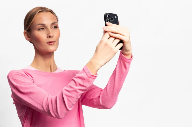 Young woman in pink shirt takes selfie with smartphone on white background. Portrait of confident female holding phone for photo, concept of social media, communication, and modern technology.