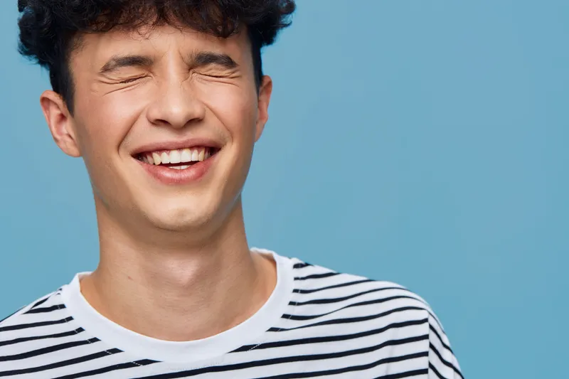 smiling man with closed eyes wearing a striped shirt on blue background expressing happiness and joy in casual style and bright lighting