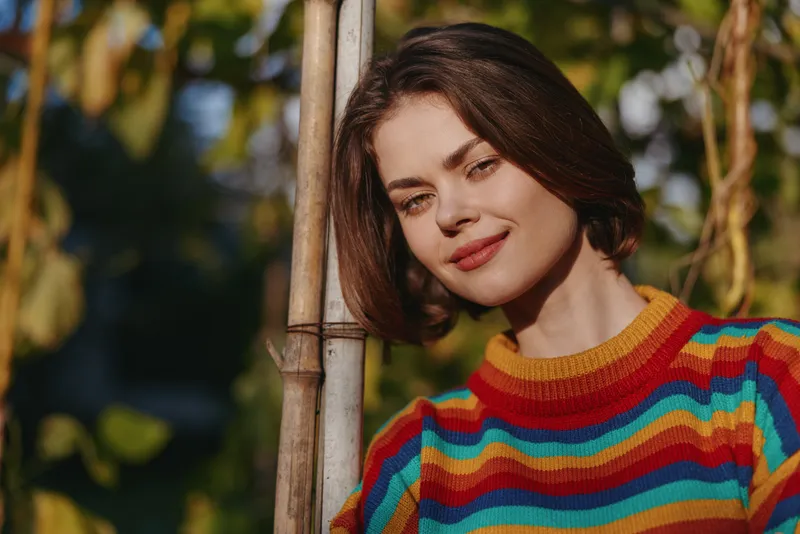 young woman striped sweater smile leaning garden outdoor portrait of early twenties female model in colorful rainbow knit, short bob hair, soft smile, relaxed pose in warm sunlight.