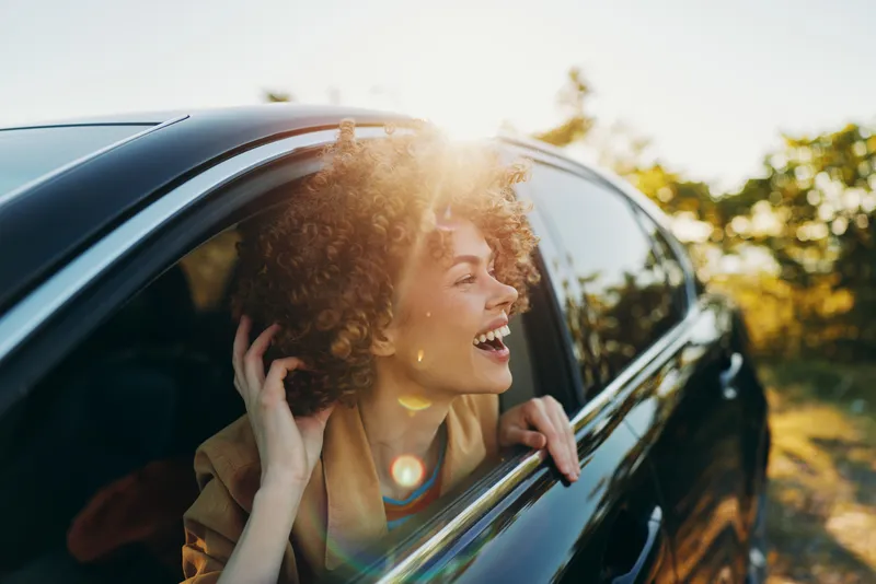 Young woman with curly hair leans out of a car window, laughing under warm sun as a retro film glow adds color to a carefree roadside moment.