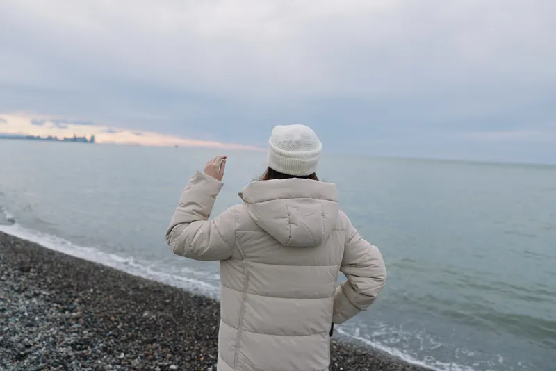 Winter scene with a person in a warm coat standing on a pebble beach, facing a calm sea and distant horizon. Cozy jacket, muted tones, serene coast atmosphere at twilight by the water.