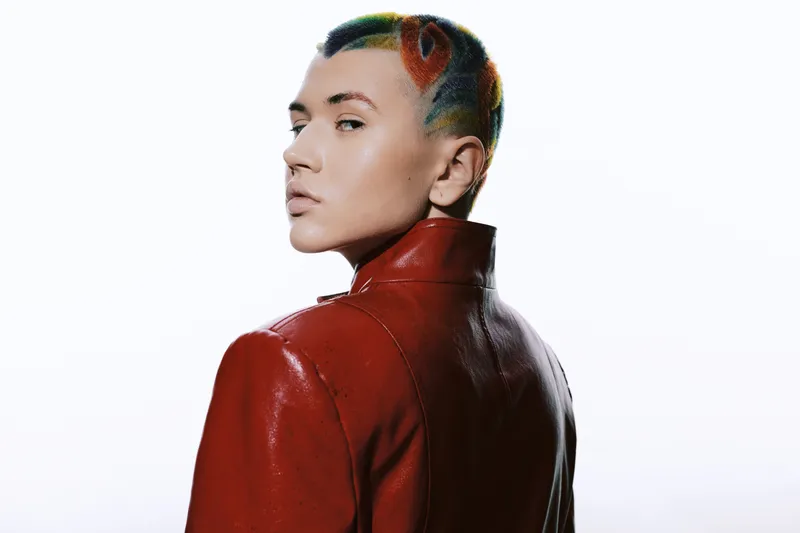 Stylish woman with short colorful hair wearing a modern red leather jacket, posing confidently against a bright white background in a contemporary fashion setting.