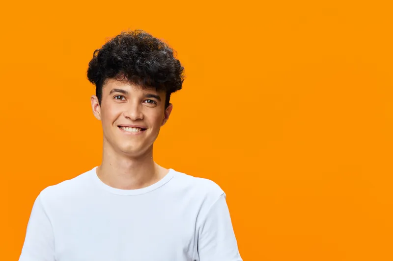 smiling man with curly hair wearing white t-shirt stands against bright orange background, expressing happiness and youthfulness with casual style and natural lighting.