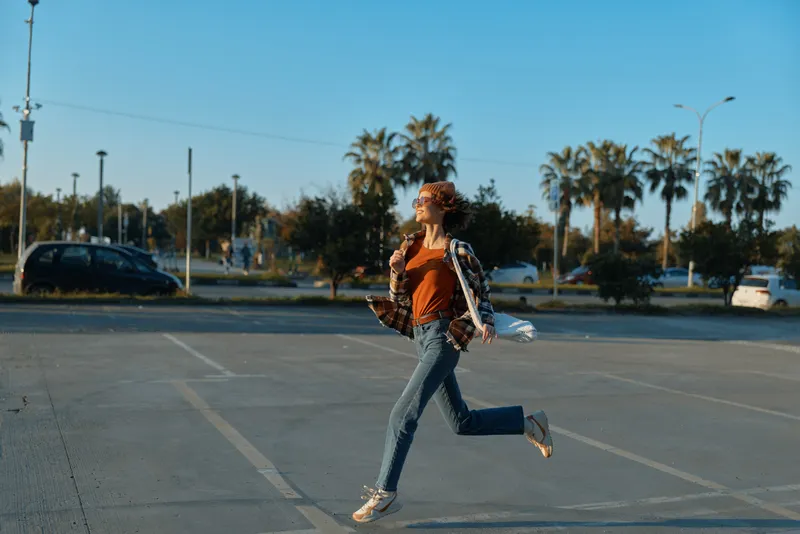 Woman jump joy urban city parking lot at golden hour glow, candid lifestyle portrait of authenticity, wearing jeans and sunglasses; emotional storytelling, mindful living and carefree modern energy.