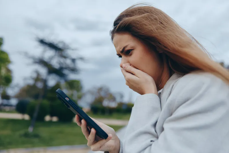 Worried woman looking at phone screen outdoors with anxious expression, emotional stress, casual clothing, cloudy sky background, financial crisis concept