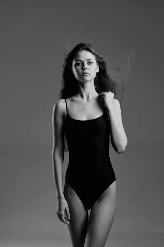 Black and white portrait of a young woman in a black bodysuit with natural expression and flowing hair in studio lighting.