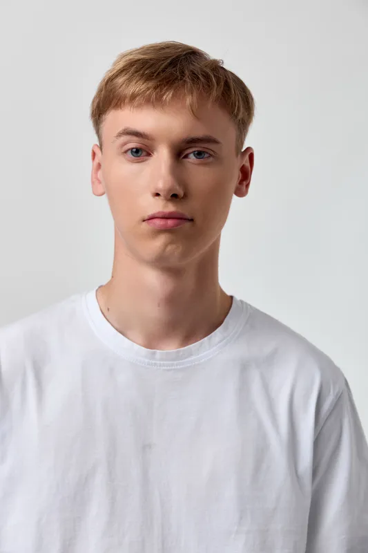 Young man in a plain white shirt stands against a neutral studio backdrop, calm and confident, ideal for fashion, lifestyle or headshot photography with clean, distraction free styling