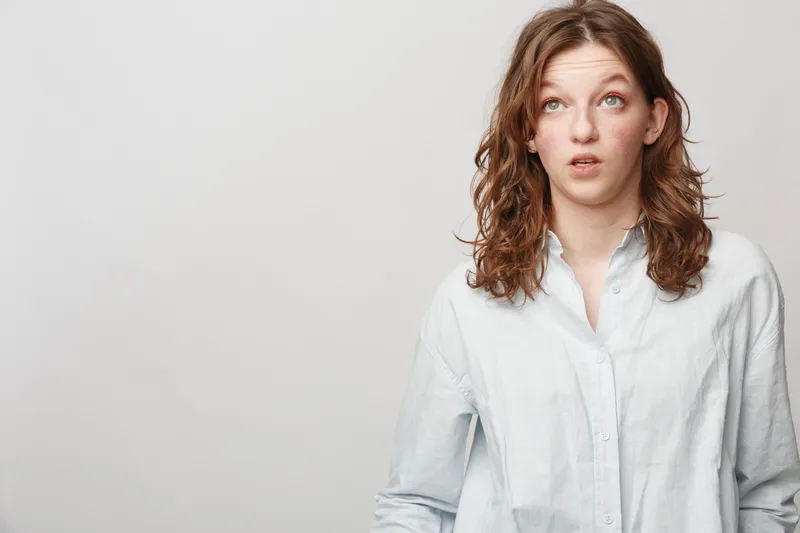 Young woman with curly hair wearing white shirt looks confused and thoughtful against plain background. Concept of doubt, uncertainty, and contemplation in casual portrait photography.