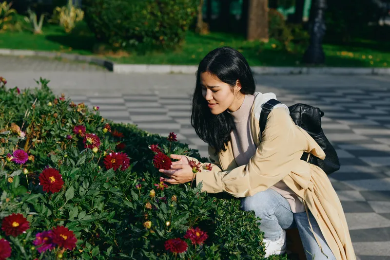 Woman kneels beside vibrant flowers in a garden, touching petals and admiring blooms in a calm outdoor scene, highlighting nature, gardening, and a peaceful daily moment in soft daylight