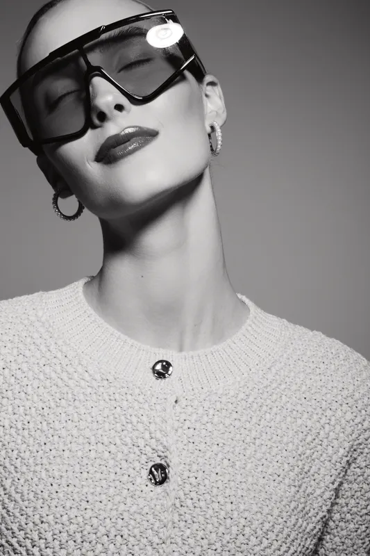 Stylish caucasian woman with pale skin wearing large sunglasses and textured sweater poses with closed eyes in black and white portrait.