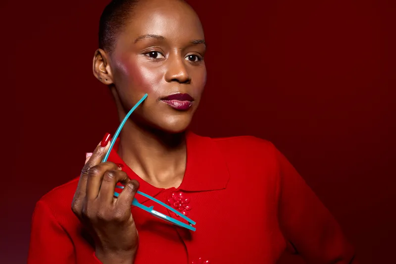 Confident African woman with dark skin and short hair holds turquoise glasses near face wearing a red sweater on red background. Concept of fashion, beauty, and style portrait studio.