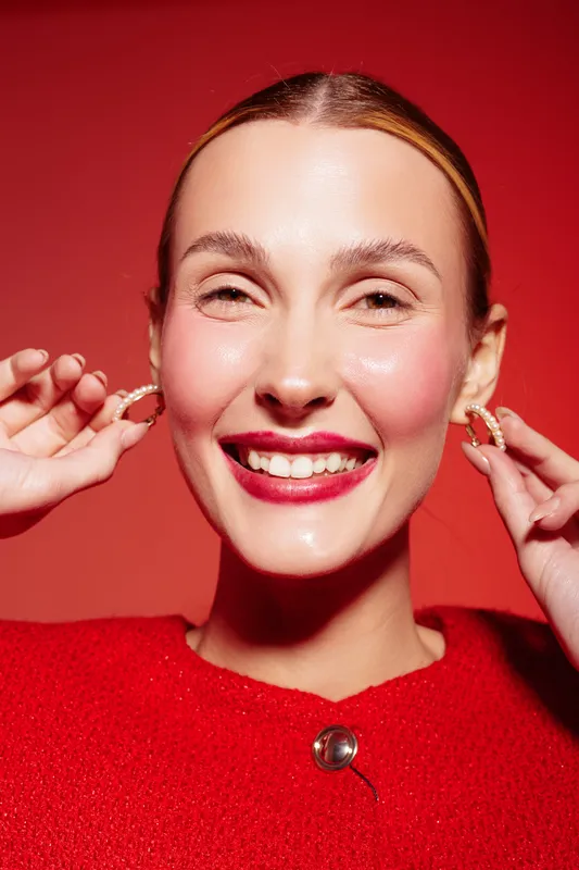 Smiling woman with red lipstick and slicked hair adjusts hoop earrings on red background. Close-up portrait with bright makeup and joyful expression. Concept of beauty and fashion.