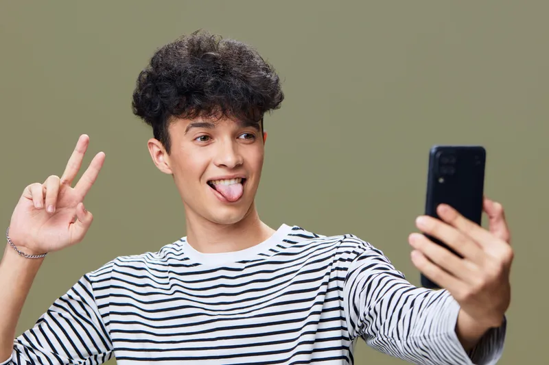 young man taking selfie with smartphone, playful expression, striped shirt, casual style, isolated background