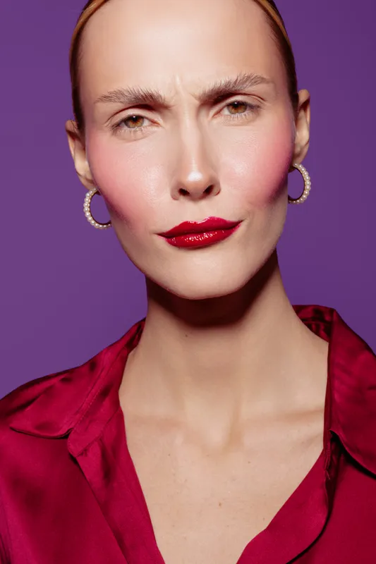 Close-up portrait of confident woman with red lipstick and hoop earrings wearing red blouse on purple background. Concept of fashion, beauty, makeup, and style in studio lighting.