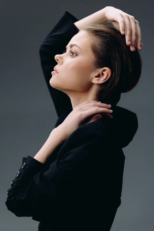 Elegant woman posing in black jacket with thoughtful expression against gray background, showcasing style and sophistication.