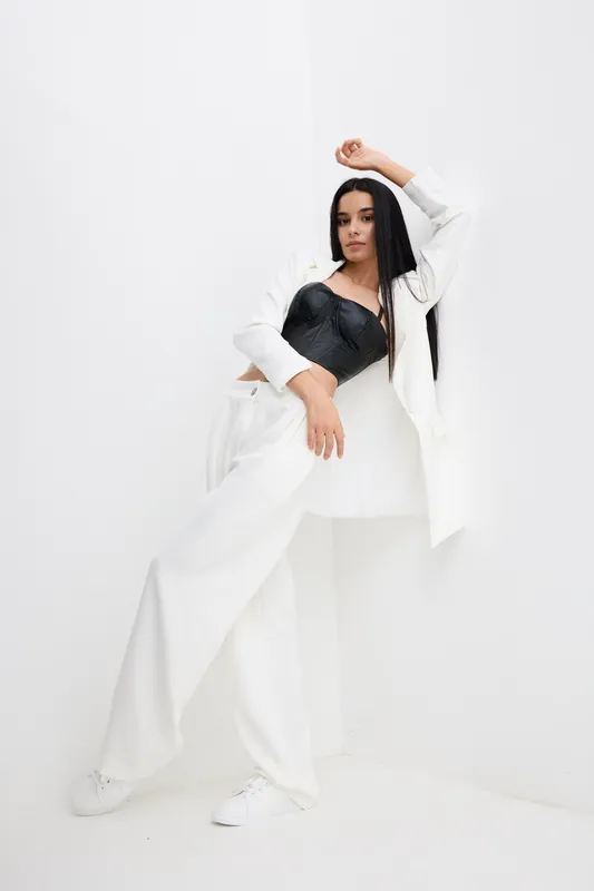 stylish woman in white wide-leg pants and jacket poses against white wall, wearing black leather corset top, long dark hair, modern fashion look, minimalistic background