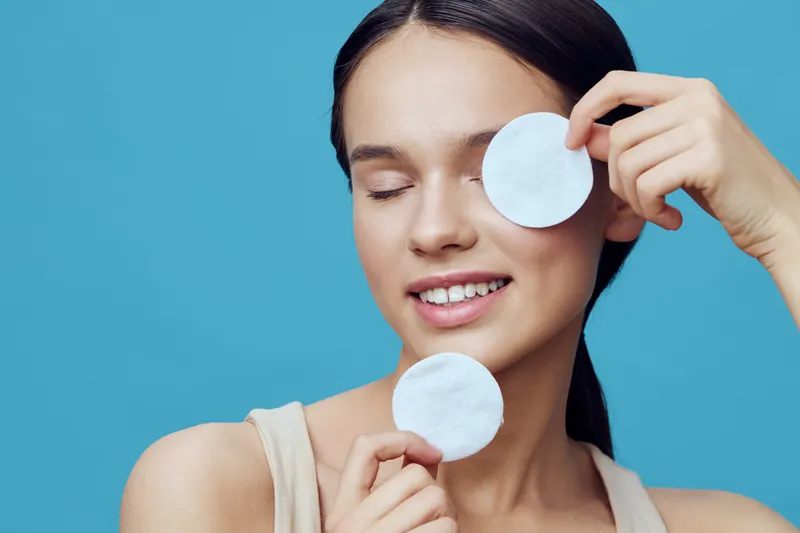 Young woman with smooth skin smiling while holding cotton pads against a blue background, representing skincare routine and beauty concept