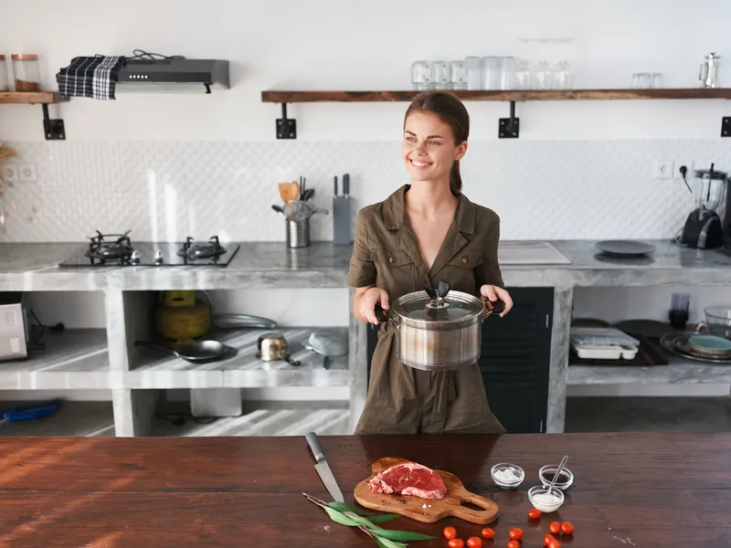 Женщина на кухне с кастрюлей в руках улыбается, готовит ужин из мяса и овощей на деревянном столе на фоне стильной кухни