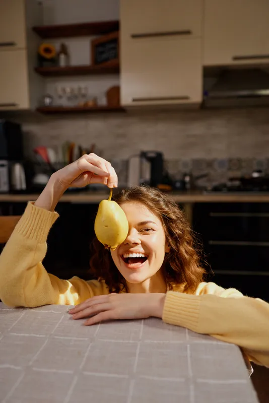 Joyful woman indoors in a cozy kitchen, smiling with a pear held overhead, relaxed posture, warm natural light, home lifestyle moment, candid and friendly