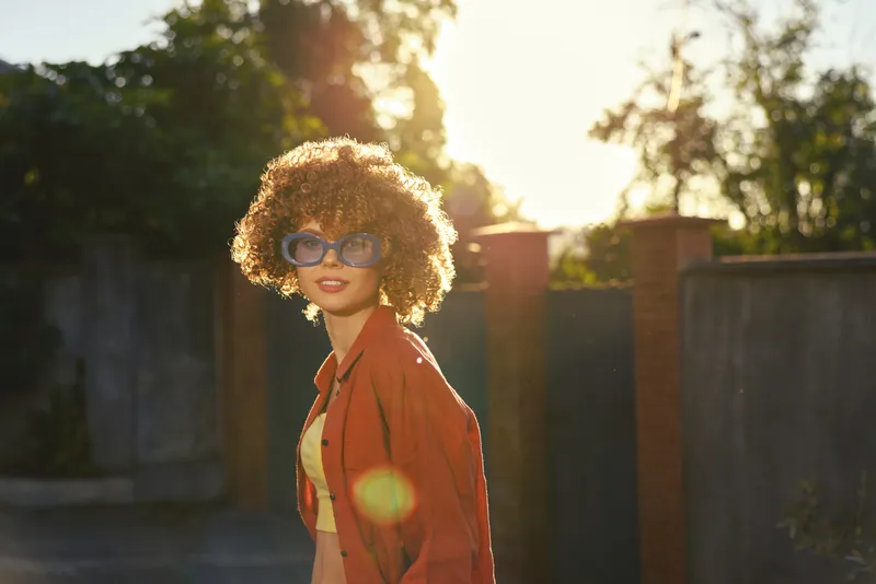 woman lifestyle portrait outdoors during sunset, wearing orange jacket and sunglasses, smiling softly, warm glow, candid street mood, urban background, relaxed pose, natural light, fresh colors