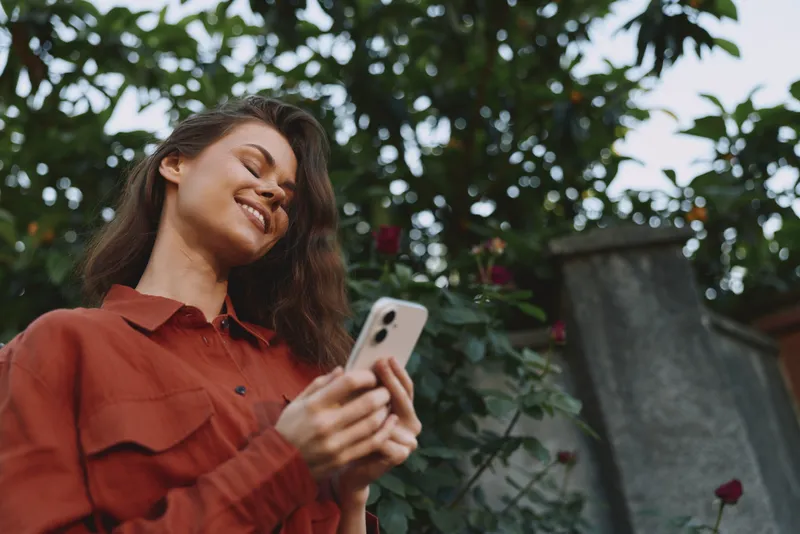 A smiling woman in a red shirt stands outdoors using a smartphone, capturing a candid moment in a casual lifestyle portrait, natural light, everyday joy