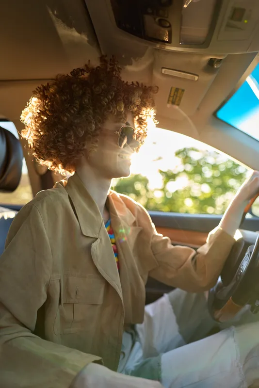Young woman with curly hair drives a car, wearing a beige blazer and sunglasses, enjoying a bright casual ride through the afternoon sun, her relaxed expression conveying a confident, free spirit