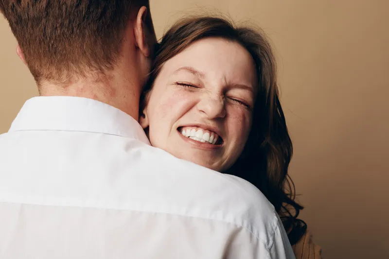 Happy woman with closed eyes smiles warmly while hugging man in white shirt. Intimate moment of affection and love captured on neutral background. Concept of relationship and joy.