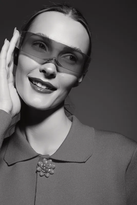 Young white woman with light skin wearing oversized translucent sunglasses and a textured sweater smiles while posing in a studio with soft lighting and monochrome effect.