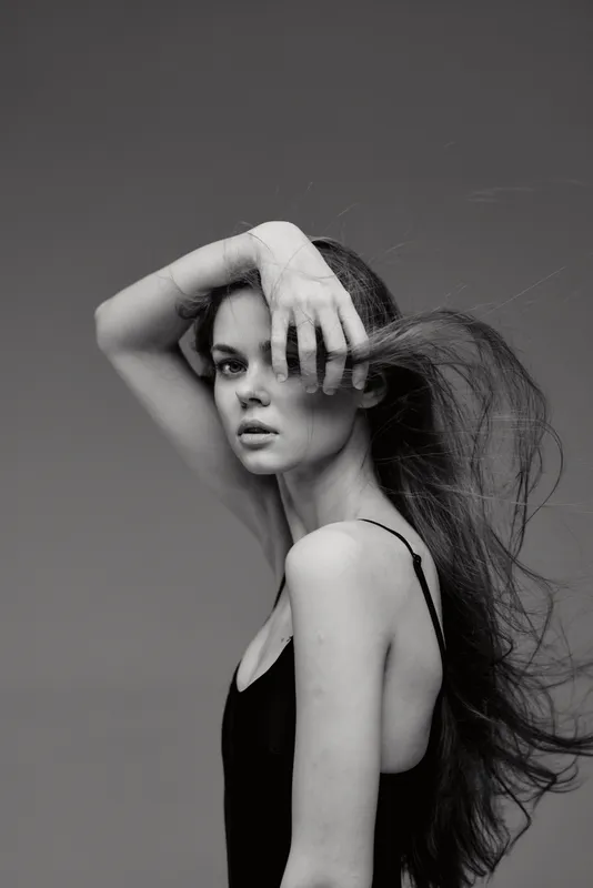 Black and white portrait of a thoughtful woman with flowing hair posing with her hand on her face, showcasing elegance and emotion in a minimalistic style.