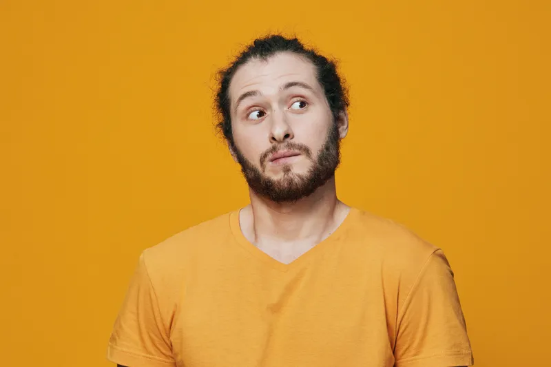 A young man with curly hair and a beard wearing a vibrant yellow t shirt, gazing curiously against a bright yellow background, expressing intrigue and playfulness