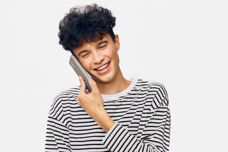 smiling man holding phone, casual young male with curly hair wearing striped shirt, happy expression, communicating, isolated white background