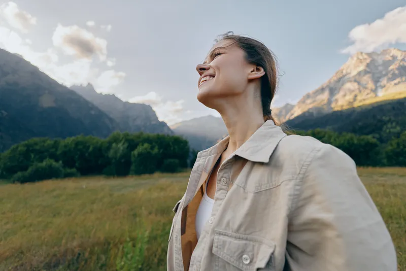 Cheerful woman outdoors, smiling with eyes closed, sunlit moment in a grassy field, mountains in the background, relaxed posture, natural colors, lifestyle vibe