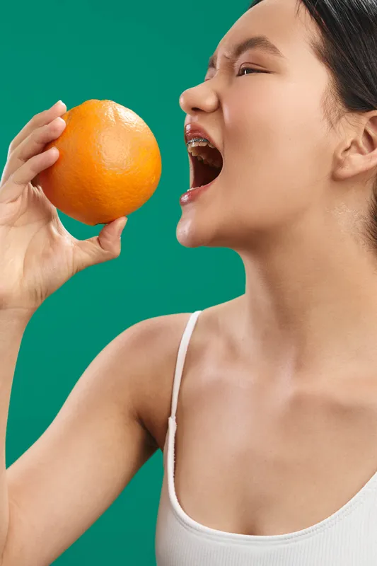 Asian woman bites into an orange against a bold green backdrop, capturing a fresh, playful moment that conveys energy, health, and vibrant style for a modern lifestyle image.