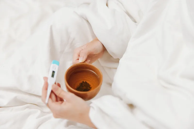 Человек держит градусник в кровати с чашкой чая, укрывшись белой простынёй поверх одеяла