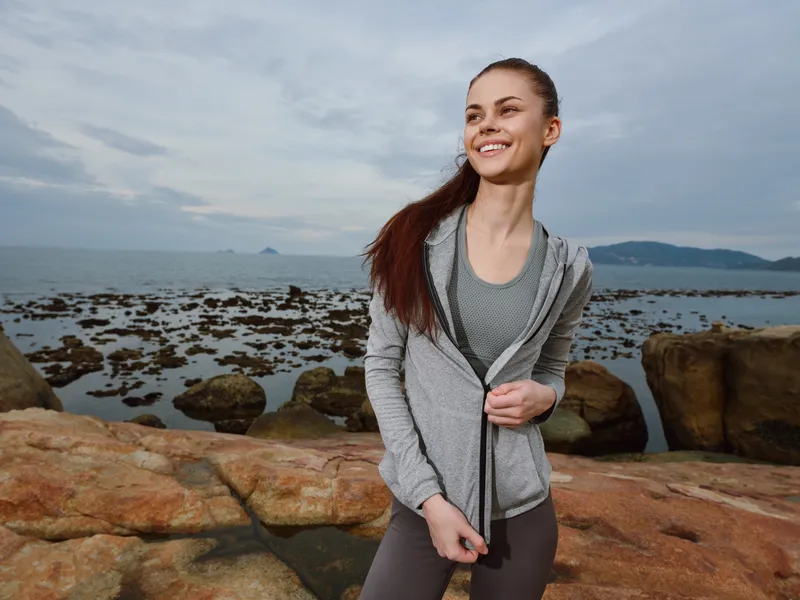 Young woman in athletic wear smiling by the ocean at sunset, showcasing a healthy lifestyle and outdoor fitness