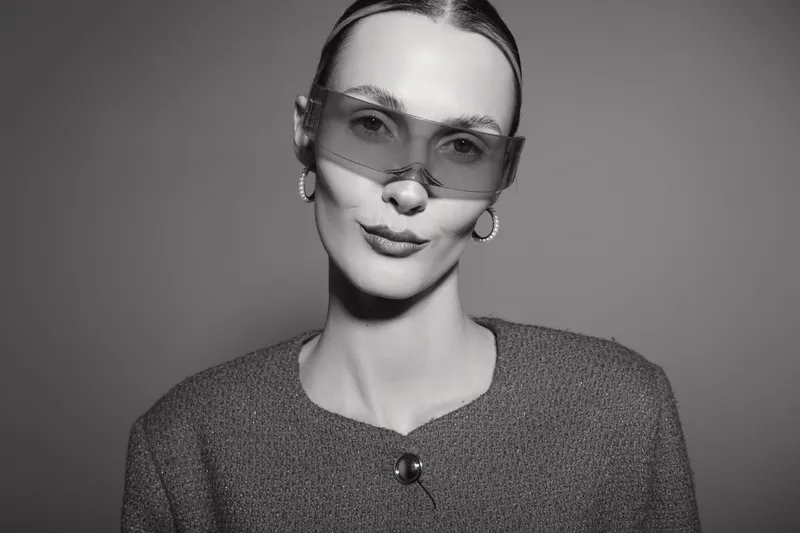Stylish caucasian woman with pale skin wears oversized sunglasses and textured jacket posing confidently in black and white studio photo. Concept of fashion, elegance and modern beauty.