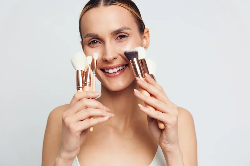 Smiling woman holding makeup brushes close to her face, clear skin, natural light, white background, beauty concept, skincare routine, fresh and happy expression