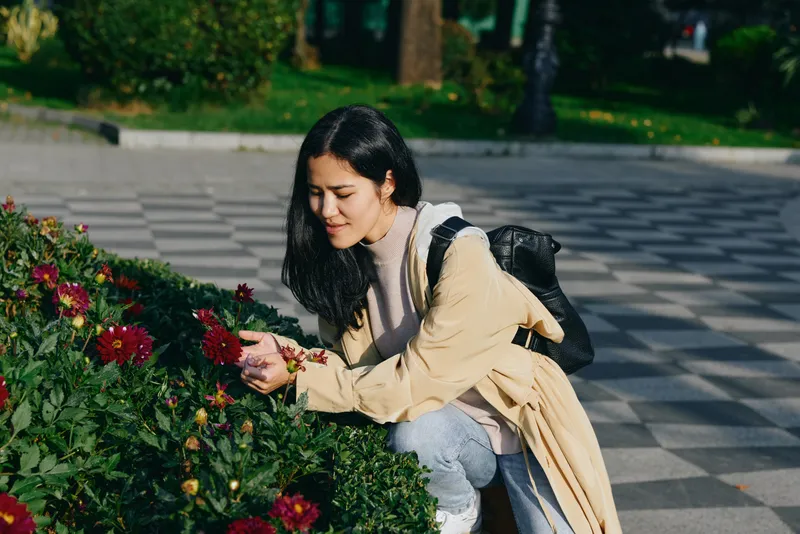 Woman crouching beside blooming flowers in a sunlit garden, admiring petals in calm outdoor scene, wearing a light coat and backpack, casual street wardrobe, capturing a peaceful moment among