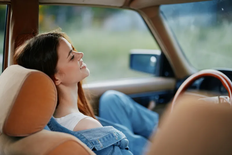 A young woman lounging in a vintage car, enjoying a sunny day outdoors, with a relaxed emotional expression and casual denim outfit.