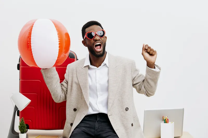 Joyful young man with dark skin, stylish glasses, and beige blazer celebrating at work desk with a beach ball on white background, emphasizing happiness and summer fun. People lifestyle concept, posit