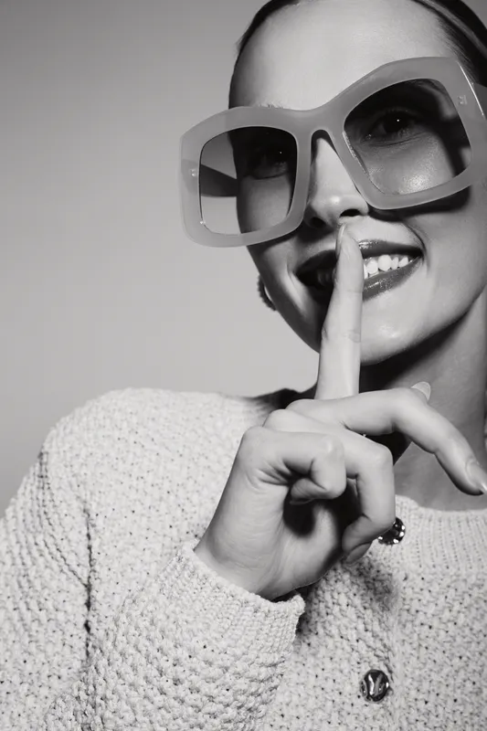 Happy Caucasian woman with fair skin wearing large sunglasses shows silence gesture with finger near lips in studio. Concept of secrecy and quiet mood.