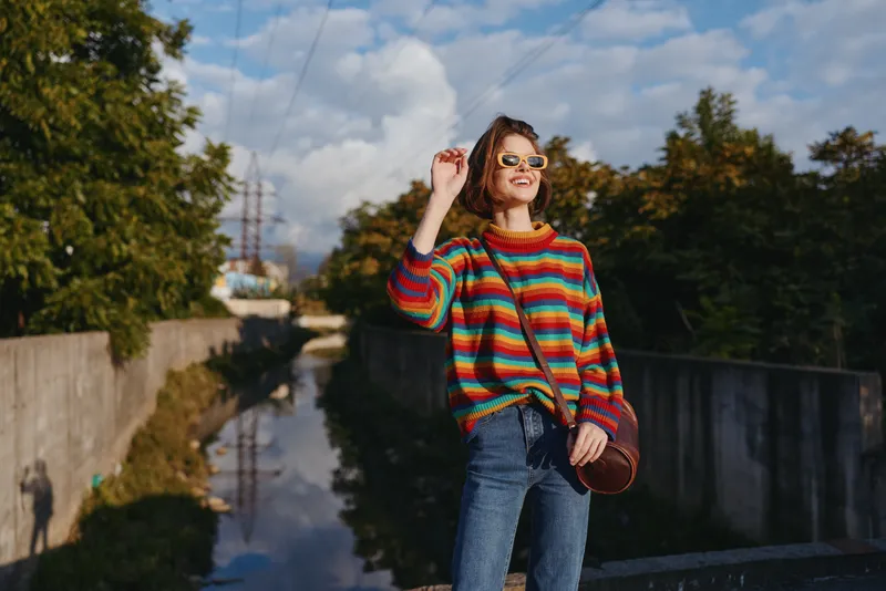 Woman in a colorful striped sweater and sunglasses, smiling by an urban canal. Young adult in jeans with crossbody bag, casual outfit, autumn trees, travel lifestyle, relaxed mood.