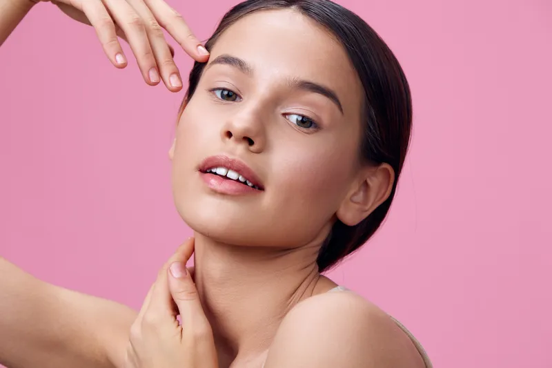 Young woman with natural beauty poses against a pink background, showcasing healthy skin and a serene expression, ideal for beauty and skincare concepts